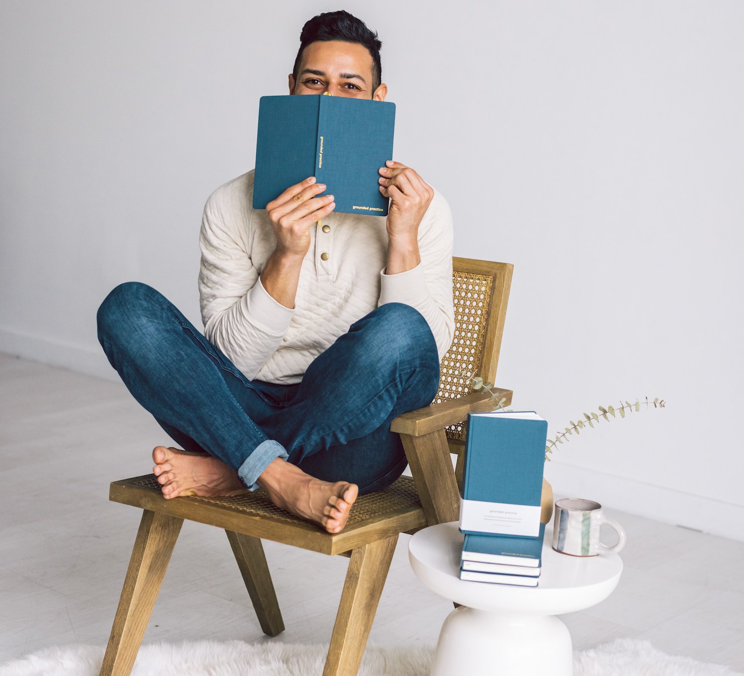 Hispanic man sitting on a woven chic boho chair covering his face with a blue linen sustainable journal and a table that has a cup of coffee and more manifestation journals. 